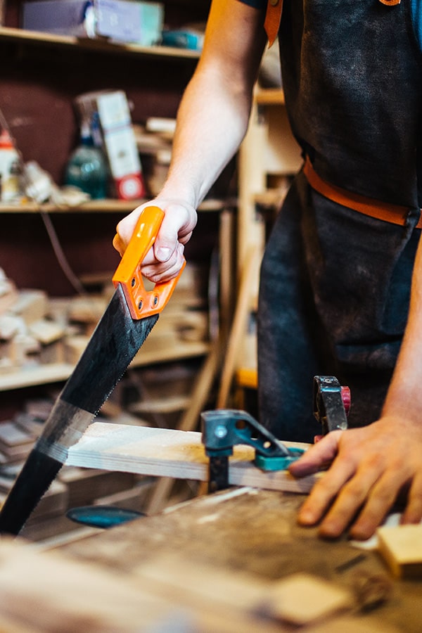 Wood worker in apron cutting a wooden board with a panel saw.